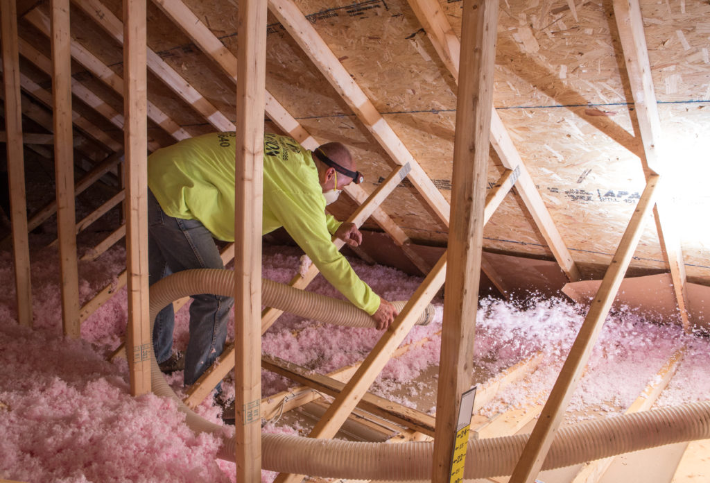 Technician blowing pink loose-fill fiberglass insulation into an unfinished attic.