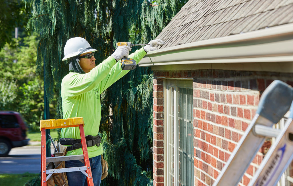 Gutter and gutter guards being installed on a brick home by a technician standing on a ladder, wearing a hard hat.