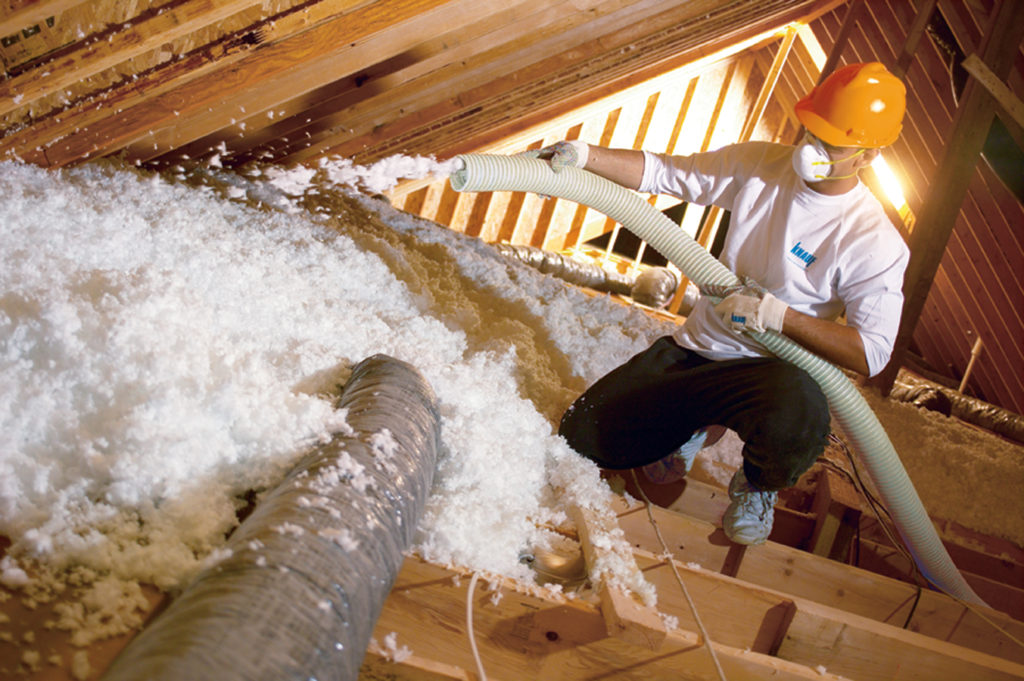 Attic loose-fill fiberglass insulation being installed by a technician wearing a yellow hardhat.